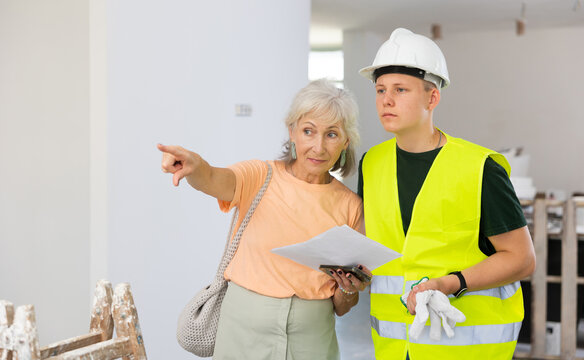 Mature woman discussing room renovation work plan with young guy in protective helmet anf yellow vest
