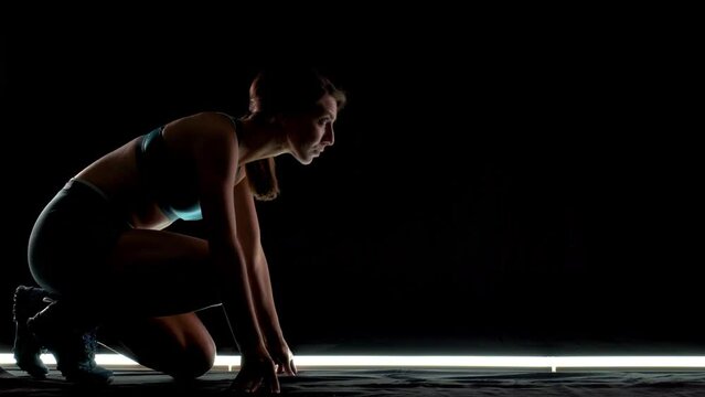 Silhouette female runner in race  start position. Girl in sportswear posing on lit track against dark background.