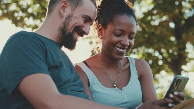 Happy smiling interracial couple watching video on mobile phone while sitting on bench