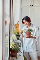 Smiling stylish Asian guy holding and caring for the plant at his flat
