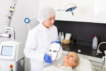 Focused elderly cosmetologist performing anti-aging face phototherapy procedure for female client in clinic of aesthetic medicine