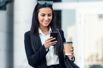 Shot of pretty young woman using her smartphone while holding a cup of coffee in the street.