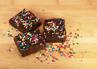 Freshly baked brownies with chocolate frosting and multicolored sprinkles are displayed on a wooden...