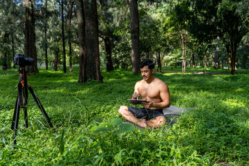 Fototapeta premium Young man practicing yoga asana, balance, meditating while standing on one leg on sports mat on green grass in park. Using tablet for online class.