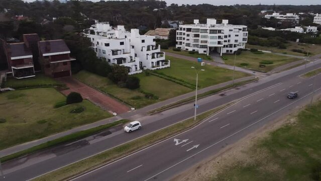 Aerial tracking shot of white car driving on boulevard road in Punta del Este, Uruguay - Luxury modern apartment buildings beside beach 