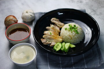 Hainanese chicken rice served on a black plate with dipping sauce and hot broth on white table.