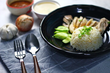 Hainanese chicken rice served on a black plate with dipping sauce and hot broth on white table.