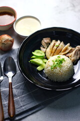 Hainanese chicken rice served on a black plate with dipping sauce and hot broth on white table.