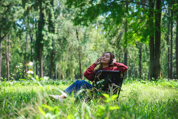 Naklejka na ściany i meble Portrait of a beautiful young asian woman enjoy listening to music with headphone while sitting on a camping chair in the park