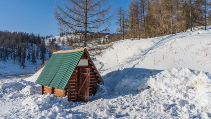 A small log cabin stands in the snowdrifts. Green triangular roof. The door is open. The forest grows on snow-covered hills. Blue sky. Altai