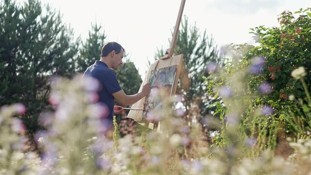 A male artist in nature paints a picture surrounded by flowers. A man paints with a brush on an easel, creativity and art. High quality 4k footage