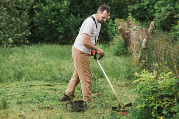 A bearded man is working in the garden. A worker mows the grass with an electric trimmer. Gardener taking care of the lawn - obrazy, fototapety, plakaty