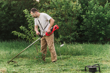 A bearded man is working in the garden. A worker mows the grass with an electric trimmer. Gardener taking care of the lawn