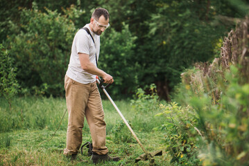 A bearded man is working in the garden. A worker mows the grass with an electric trimmer. Gardener taking care of the lawn - obrazy, fototapety, plakaty