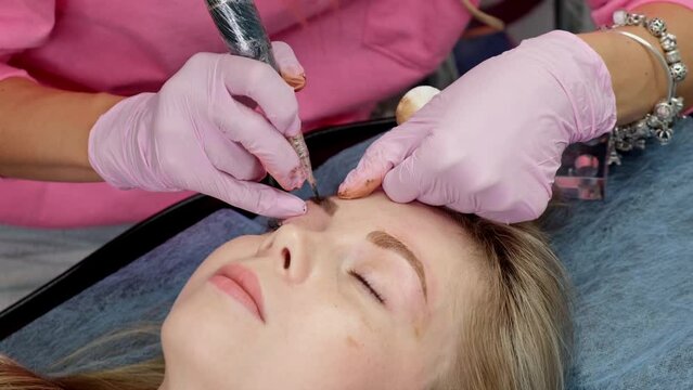 Young woman receiving permanent make up in beauty salon. Eyebrow microblading is performed using manipulator handle and special nozzle with needles. Permanent make-up und microblading.