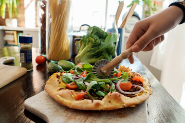Shot of woman cutting home made pizza preparing it for serving in daytime at home.