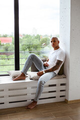 Barefoot african american man looking at camera while sitting on windowsill at home 