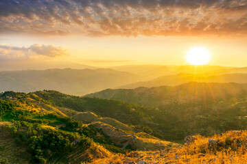 Fototapeta premium Mountain valley during sunset or sunrise. Rocks and slopes covered with green trees with amazing cloudy sunny sky on background.