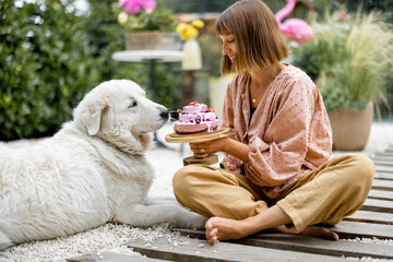 Cheerful woman playing with her adorable white dog, while sitting with festive cake in garden. Holiday celebration with pets concept