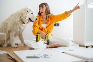 Young woman assembles furniture by herself, sitting and having fun with her cute dog at new apartment. DIY and house improvement concept