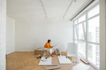 Young woman assembles furniture by herself, sitting with dog near window and reading manual at new apartment. Wide view with copy space