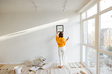 Young woman hanging picture frame in room, decorating her newly renovated apartment, stands with...