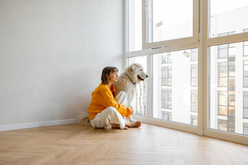 Young woman sits with her dog near the window in bright room of her new apartment. Friendship with...