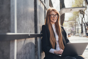 Portrait of pleased red-headed woman in eyeglasses sitting on bench in park and working on laptop computer. Online work on the Internet. Distance learning - Powered by Adobe