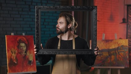 Positive caucasian male artist smiling cheerful on camera through black vintage picture frame. Portrait of happy bearded man in apron posing in creative work space.