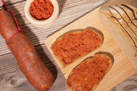 Sobrasada de Mallorca. Dos rebanadas de pan moreno con sobrasada d e Mallorca, junto a una sobrasada y un cuenco con piment&oacute;n, sobre una tabla de madera.