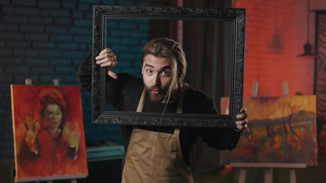 Handsome caucasian man looking through black vintage frame with curiosity on face. Crazy talented artist in apron working on own modern art studio.