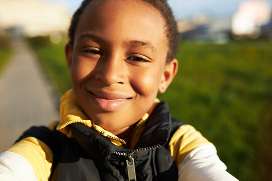 Handsome smiling kid boy of black ethnicity taking selfie on front camera of his smartphone, happy with new mobile phone, pretending to be blogger, walking down city streets or park on sunny day