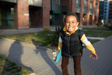 Black boy kid with skateboard in hands roaring at camera showing teeth on city street background,...