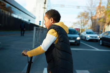 Side view of african american boy kid crossing street on scooter in traffic jam, going to skate...