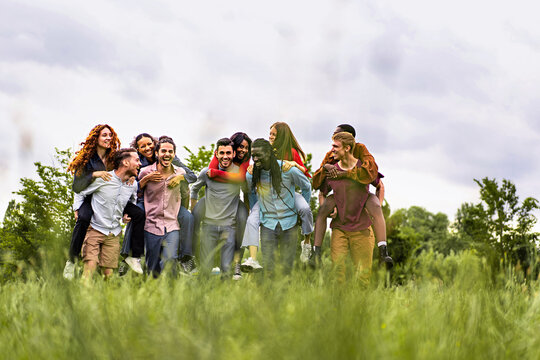 Group of multiethnic young cheerful people walking together and playing piggyback in the countryside - happy friends having fun gathering in the weekend - trust, unity and diversity lifestyle concept