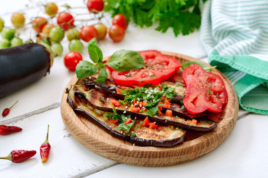 Grilled eggplant slices with spicy seasoning and greens, fresh tomato on a wooden plate. Vegetarian dish