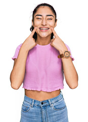 Hispanic teenager girl with dental braces wearing casual clothes smiling with open mouth, fingers...