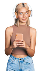 Beautiful young blonde woman using smartphone wearing headphones smiling looking to the side and...