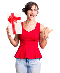 Beautiful young woman with short hair holding gift pointing thumb up to the side smiling happy with open mouth