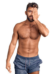 Fototapeta premium Young hispanic man wearing swimwear shirtless covering one eye with hand, confident smile on face and surprise emotion.