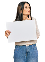 Middle age brunette woman holding blank empty banner serious face thinking about question with hand on chin, thoughtful about confusing idea