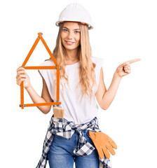 Beautiful caucasian woman with blonde hair wearing architect hardhat and holding tools smiling...