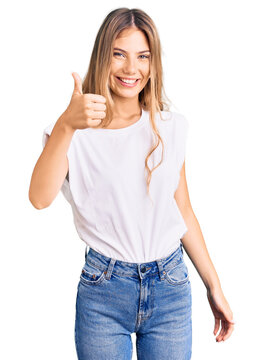 Beautiful caucasian woman with blonde hair wearing casual white tshirt doing happy thumbs up gesture with hand. approving expression looking at the camera showing success.