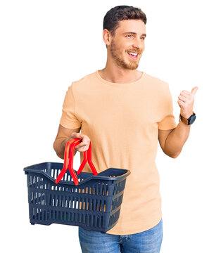 Handsome young man with bear holding supermarket shopping basket pointing thumb up to the side smiling happy with open mouth