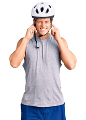 Young handsome man wearing bike helmet covering ears with fingers with annoyed expression for the noise of loud music. deaf concept.