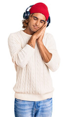 Young hispanic man listening to music using headphones sleeping tired dreaming and posing with hands together while smiling with closed eyes.
