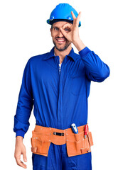 Young handsome man wearing worker uniform and hardhat doing ok gesture with hand smiling, eye looking through fingers with happy face.