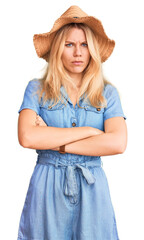 Young beautiful blonde woman wearing summer hat and dress skeptic and nervous, disapproving expression on face with crossed arms. negative person.