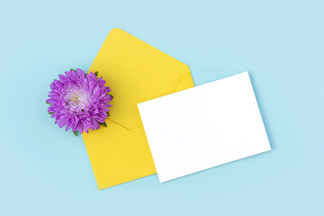 White blank card, yellow envelope and purple flower on blue background. Minimal style. Top view Flat lay Mockup