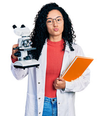 Young hispanic woman with curly hair holding microscope and book looking at the camera blowing a...